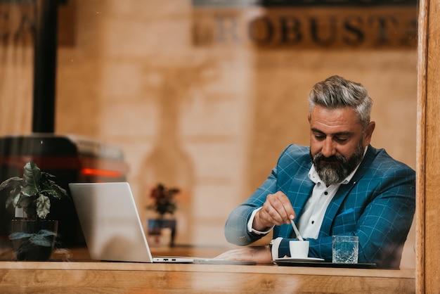 Modern senior businessman sitting in cafe and using laptop and smartphone