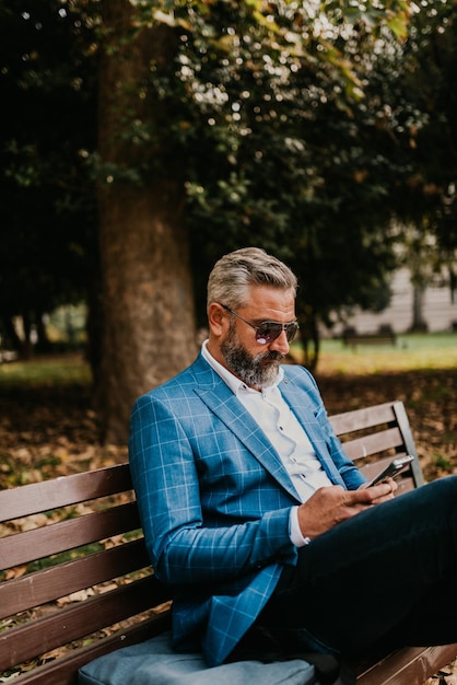 Modern senior businessman using smartphone for online meeting while sitting on bench