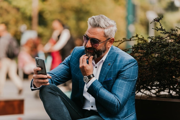 Modern senior businessman using smartphone for online meeting while sitting on bench