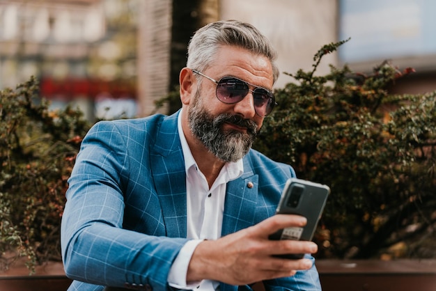 Modern senior businessman using smartphone for online meeting while sitting on bench