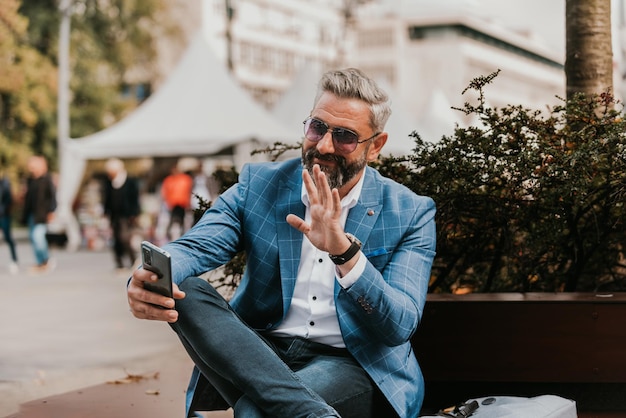 Modern senior businessman using smartphone for online meeting while sitting on bench