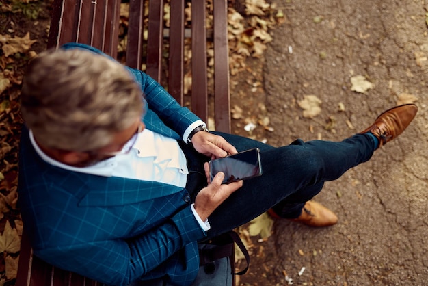 Modern senior businessman using smartphone for online meeting while sitting on bench .