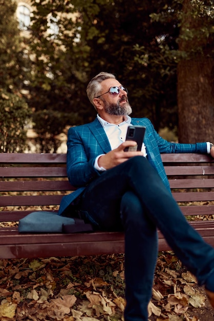 Modern senior businessman using smartphone for online meeting while sitting on bench .