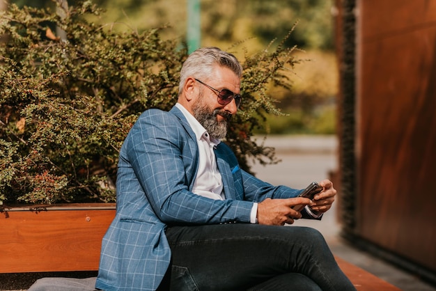 Modern senior businessman using smartphone for online meeting while sitting on bench.