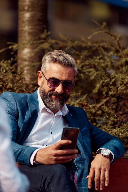 Modern senior businessman using smartphone for online meeting while sitting on bench .