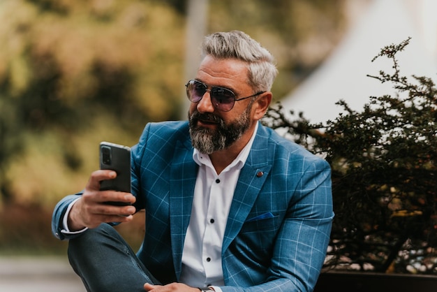 Modern senior businessman using smartphone for online meeting while sitting on bench.