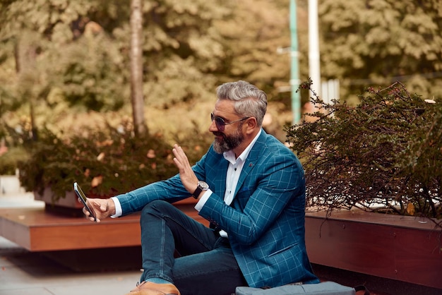 Modern senior businessman using smartphone for online meeting while sitting on bench .