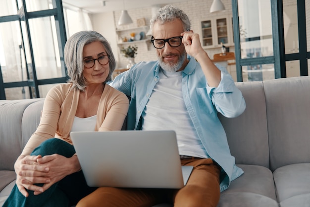 Modern senior couple in casual clothing smiling and using laptop while bonding together at home