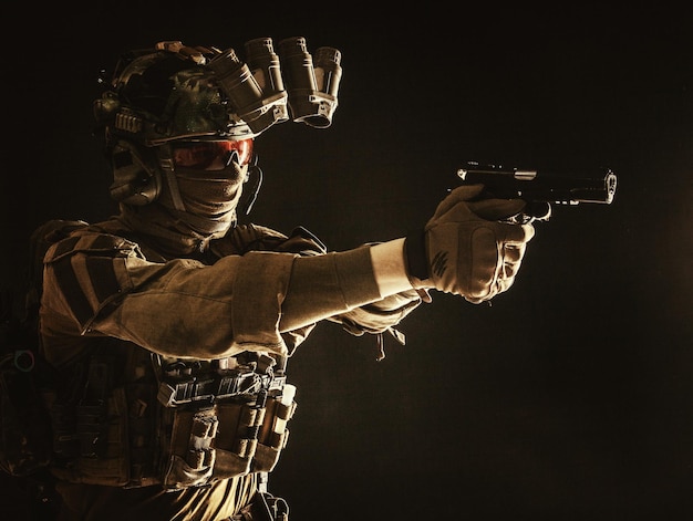 Modern soldier counter terrorist squad fighter in combat uniform helmet and tactical radio headset aiming service pistol in darkness shooting with handgun studio portrait on dark background