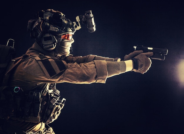 Modern soldier counter terrorist squad fighter in combat uniform helmet and tactical radio headset aiming service pistol in darkness shooting with handgun studio portrait on dark background