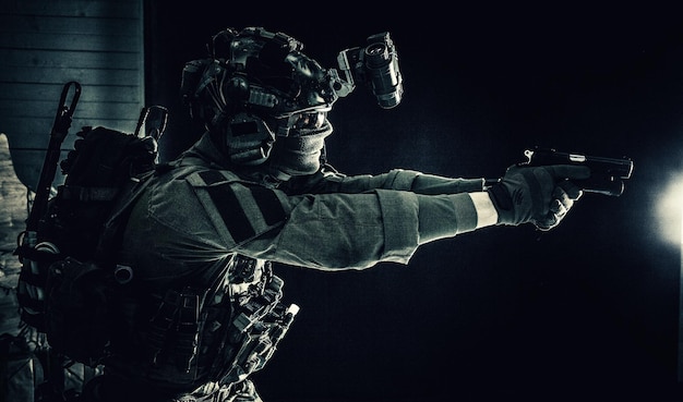 Modern soldier counter terrorist squad fighter in combat uniform helmet and tactical radio headset aiming service pistol in darkness shooting with handgun studio portrait on dark background