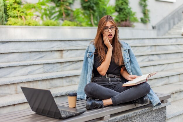 Modern students. Distance learning. Young enthusiastic woman reads book while sitting on bench with a laptop