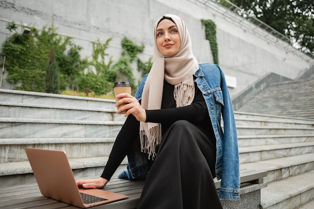 Modern stylish muslim woman in hijab, denim jacket and black abaya sitting in city street working on laptop