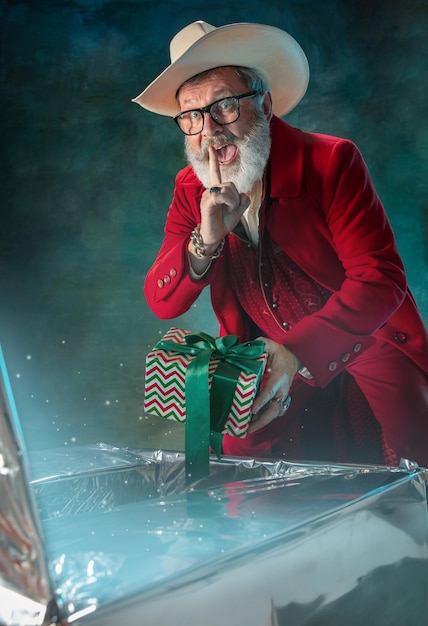 Modern stylish Santa Claus in red fashionable suit and cowboy's hat on dark