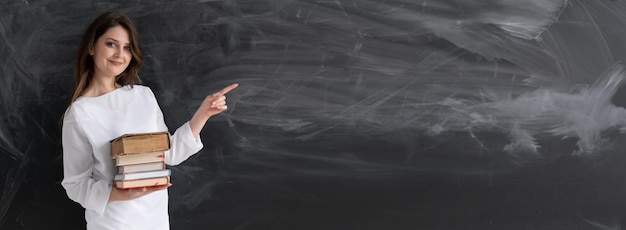 Modern teacher woman 30 years old with textbooks in her hands stands in the classroom near the blackboard with a place for the text Online lesson and teacher work School education Banner