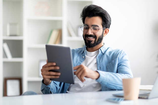 Modern technologies smiling young indian man using digital tablet at home office