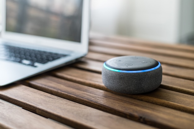 Modern wireless smart speaker placed on wooden table near blurred laptop at home