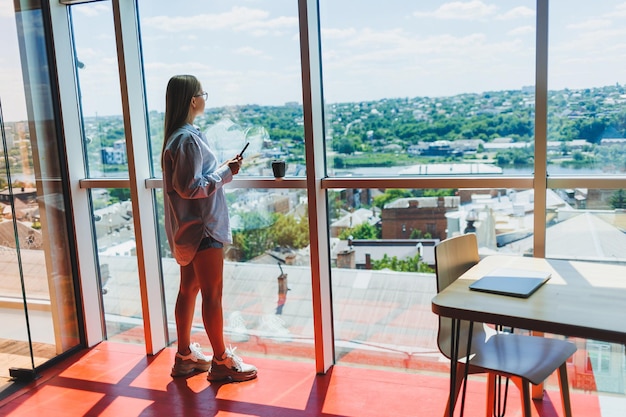 Modern woman freelancer talking on the phone while standing at a large window and drinking coffee The concept of a modern successful woman Young girl in an open office