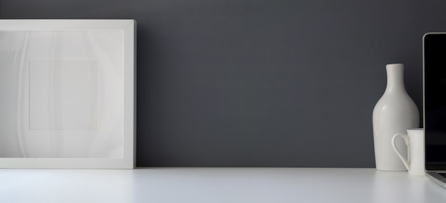 Modern workspace with blank frame and ceramic vase on white table and grey wall background 