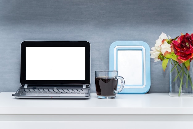 Modern workspace with blank screen laptop, frame, coffee cup and vase on white table with gray-blue wall background