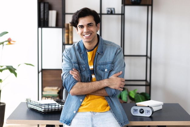 Modern young entrepreneur with arms crossed looking at camera and smiles friendly. Successful young businessman or manager stands near his workplace in the office