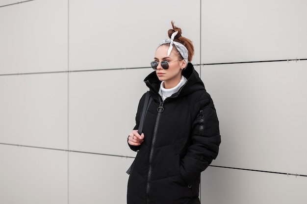 Modern young hipster woman in round sunglasses in a stylish coat in a fashionable bandana with a leather backpack is standing near a white wall a warm winter day. Fashionable american girl on vacation