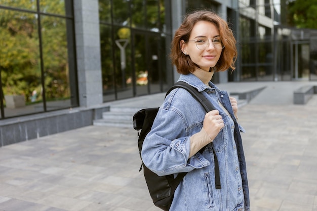 Modern young student woman in stylish denim jacket and backpack outdoors