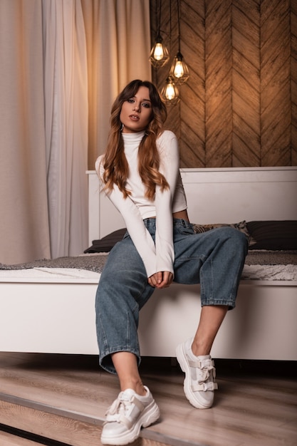 Modern young woman with long red hair in jeans in a white shirt in shoes sits on a soft bed in the room with a vintage interior. Sexy girl model in stylish clothes is resting in the bedroom. Fashion.