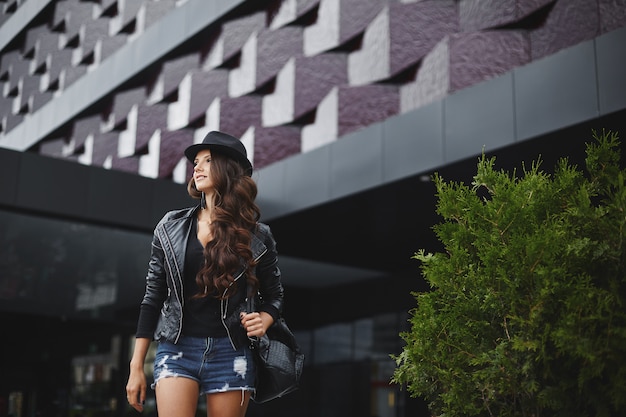 Photo modish model girl with wavy hair in a leather jacket, black hat and jeans shorts with a stylish backpack stands on the street near modern building. trendy casual outfit. everyday look. street fashion.