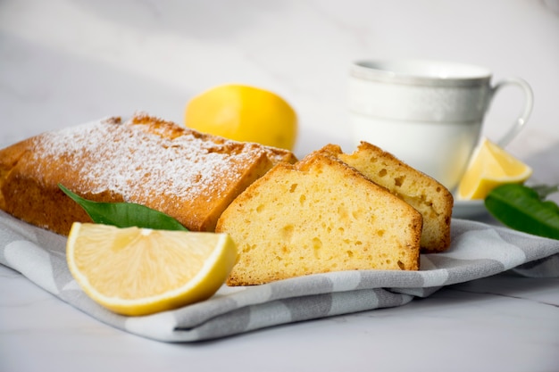 Moist lemon pound cake on kitchen towel on marble table with slices of lemon and cup of tea on plate. Delicious breakfast, traditional tea time treat. Reciepe of English lemon pie loaf.