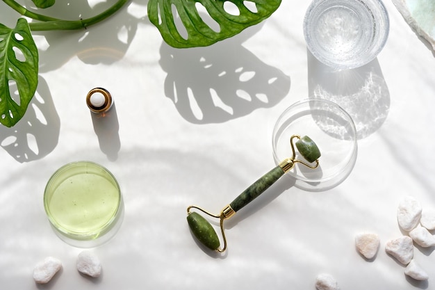 Moisturizer green jade face roller with exotic leaves Monstera Adansonii leaves Sunshine long shadows Minimal flat lay on off white background Facial massage with self made cosmetics