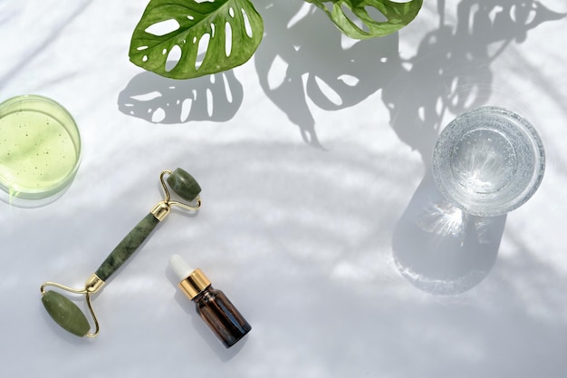 Moisturizer, green jade face roller with exotic leaves. Monstera Adansonii leaves. Sunshine, long shadows. Minimal flat lay on off white background. Facial massage with self made cosmetics.