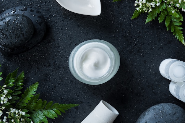 Moisturizer jar with white tubes of lotion and basalt rock on black background.