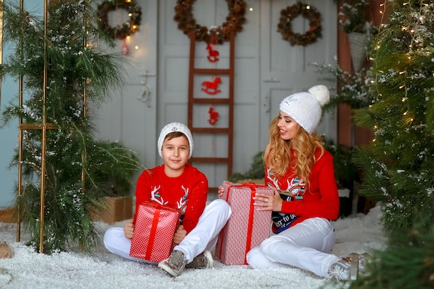 Mom and adult son sitting on the ground surrounded by artificial snow wearing hats white pants and a warm sweater.