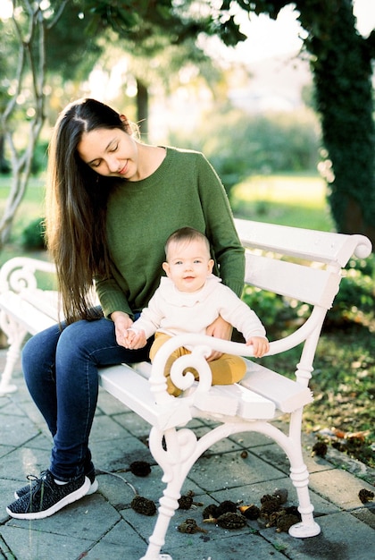 Mom and baby are sitting on a white bench in a green park