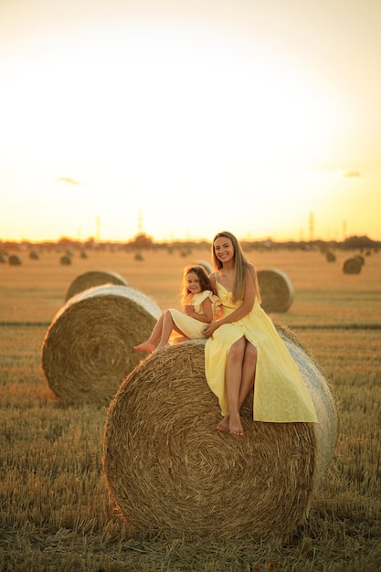 Mom and blonde daughter are sitting on a haystack in a field at sunset Mother and child have fun on the farm