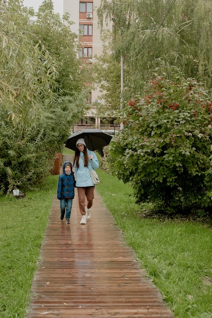 Mom in a blue jacket walks in the park with her son along a wooden path with an umbrella