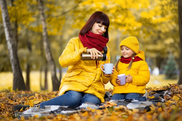 Mom and cute daughter, sitting on a blanket in beautiful golden autumn park. Pretty woman pouring hot tea from a thermos to cups