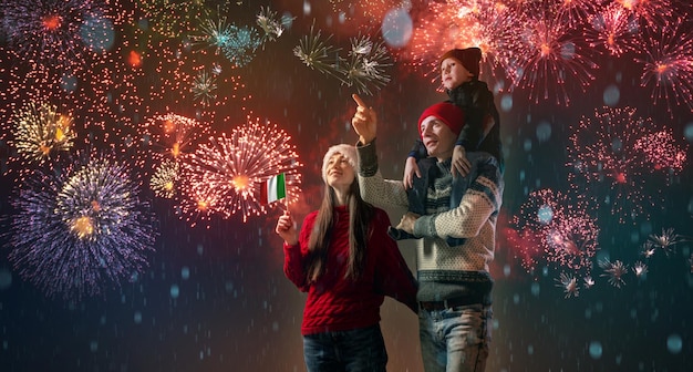 Photo mom and dad with their son on their shoulders in warm clothes and in a santa hat the family celebrates the new year looking at the fireworks outside