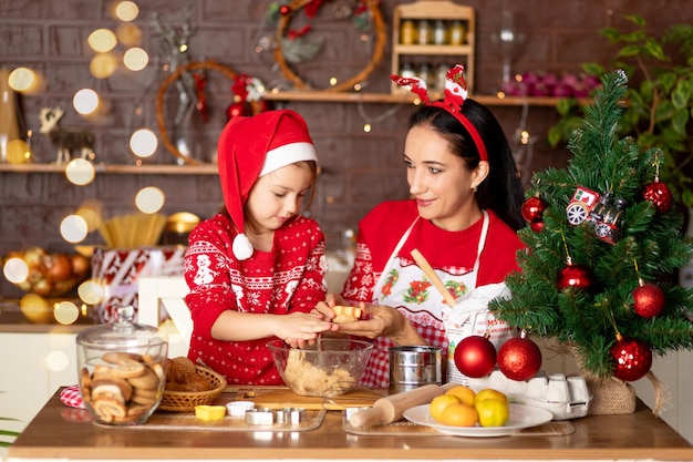 Mom and daughter are cooking ginger cookies in the kitchen and preparing for the new year and Christmas, having fun and rejoicing in red sweaters and Santa Claus hat