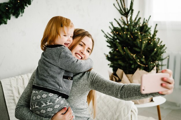 Mom and daughter are photographed on the phone near the Christmas tree. Mother and little child having fun, hugging and playing together near at home. Merry Christmas and Happy Holidays.