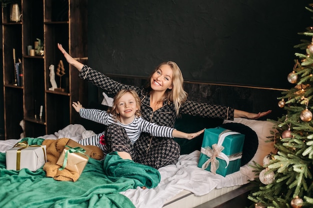 Mom and daughter are sitting in pajamas on the bed by the Christmas tree at Christmas New Year family holiday gifts around christmas family morning