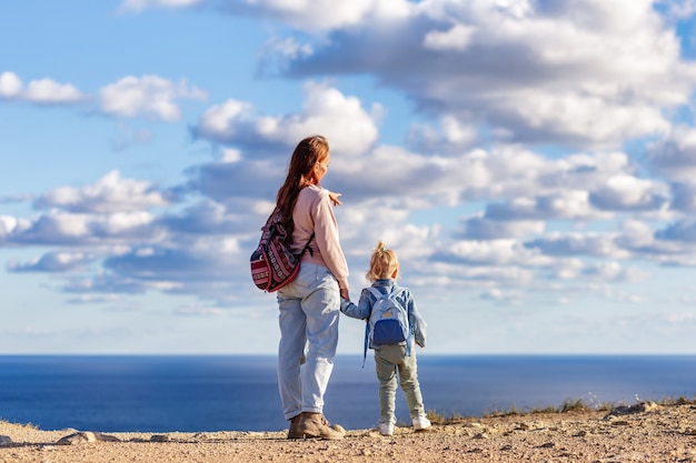 Mom and daughter have climbed a mountain and are looking at the sea with their backs to the camera