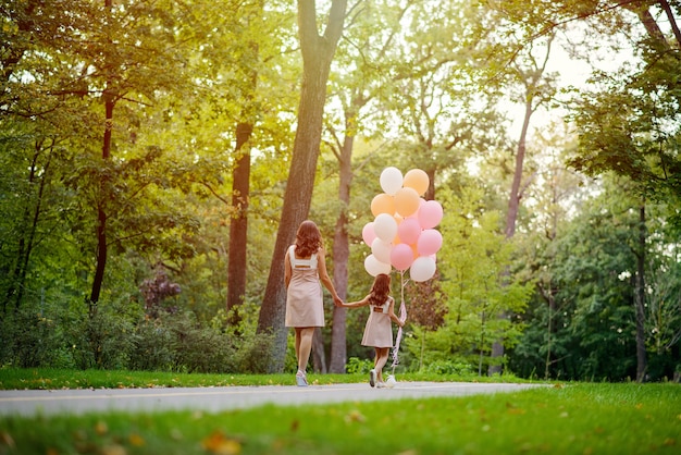 Mom and daughter holding hands walk in the summer in the park with a large bunch of balloons. Mother's love.