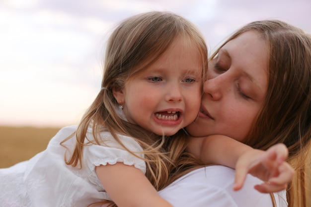 mom and daughter hug and kiss each other on the background of the sky and wheat