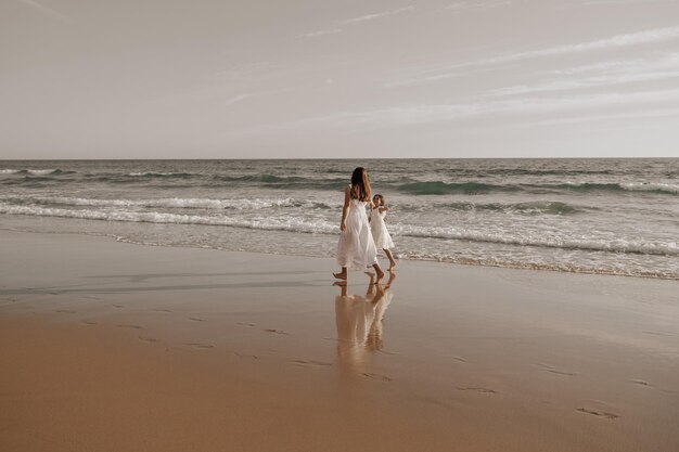 Photo mom and daughter in similar white dresses walking on sandy seashore