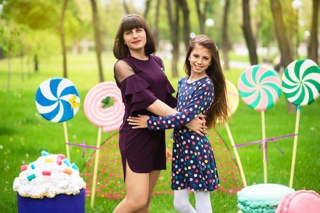 Mom and daughter standing and hugging in the scenery from huge sweets of lollipops and cakes