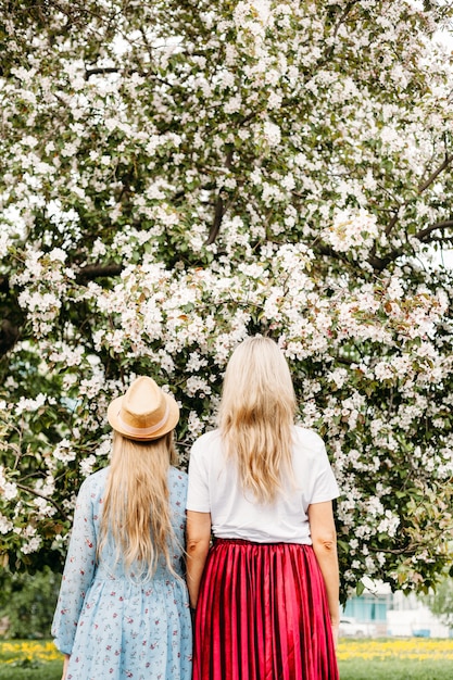 Mom and daughter teen back near a flowering apple tree, walking around the city, view behind, bright clothes