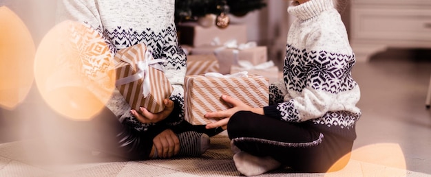 Mom and daughter in white sweaters with black patterns and black pants sit on the floor and give each other Christmas gifts. Photo