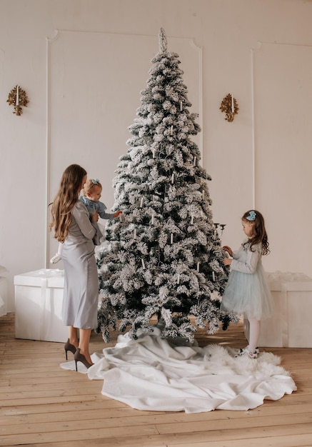 Mom and daughters in festive dresses decorate a huge Christmas tree in the room